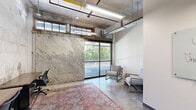 This single-floor office space blends industrial character with functional workspace design. The open-plan layout accommodates multiple workstations fitted with ergonomic task chairs and storage pedestals, all anchored by a richly patterned area rug that adds warmth to the raw aesthetic. Exposed concrete block walls, visible ductwork, and yellow-accented structural elements overhead lend the space an authentic industrial character, while polished concrete floors throughout reinforce the utilitarian yet stylish atmosphere.

A standout feature is the large glass-panel garage-style door along one wall, which floods the space with natural light and provides direct views of the surrounding greenery and outdoor area. Adjacent to this opening, a dedicated lounge nook hosts two upholstered armchairs paired with a stone side table, creating a casual meeting or relaxation zone distinct from the primary work area.

A glass whiteboard mounted on a clean white wall serves as a collaborative tool and visual anchor near the entry. A glazed interior door connects to an adjacent room visible in the background, suggesting easy access to additional workspace. Overhead fluorescent strip lighting complements the natural light, ensuring the space remains well-illuminated throughout. The overall ambiance balances creative, loft-inspired design with practical office functionality.