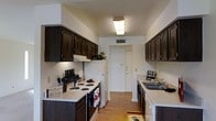 This single-floor space is thoughtfully designed to maximize functionality and comfort. The kitchen serves as a standout feature, showcasing a classic galley layout with rich, dark-stained cabinetry lining both walls from floor to ceiling, providing generous storage throughout. Light-toned countertops contrast beautifully with the cabinetry, creating a balanced and visually appealing workspace. The kitchen is equipped with a full suite of appliances, including a range with coil burners, a refrigerator, and a dishwasher, all seamlessly integrated into the layout. A double-basin sink sits beneath under-cabinet task lighting, adding both practicality and a warm ambiance. Warm hardwood-style flooring runs the length of the kitchen, adding a welcoming and cohesive aesthetic underfoot. Overhead, a flush-mount light fixture provides bright, even illumination throughout the space. A ceiling vent ensures comfortable climate control year-round. The kitchen corridor leads naturally toward a doorway, hinting at a well-connected and efficient floor plan. Overall, the space strikes a balance between practicality and inviting design, making it a comfortable and fully functional environment.
