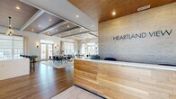 The Heartland clubhouse is a thoughtfully designed single-floor amenity space that blends contemporary aesthetics with warm, natural materials. Upon entry, a striking reception desk clad in light wood paneling greets visitors, set against a textured stone-look accent wall bearing the Heartland name. The open-concept main area seamlessly connects the kitchen and living room, anchored by wide-plank hardwood-style flooring and coffered ceilings with rich wood inlays and recessed lighting throughout.

The kitchen is outfitted with slate-blue cabinetry, white quartz countertops, stainless steel appliances, and a textured brick-style accent wall that adds warmth and character. An island provides additional counter space, while large windows flood the area with natural light and outdoor views.

The living area is a curated lounge space featuring a linear stone fireplace, a flat-screen television, bold original artwork, and statement ring-chandelier lighting. Plush seating arrangements and patterned rugs create distinct zones for relaxing and socializing. A sun-drenched reading nook with floor-to-ceiling windows offers a quieter retreat.

A private workstation with a built-in desk, large round mirror, and task lighting provides a functional workspace. A hallway connects the main areas to the full bathroom, completing this well-appointed communal facility.