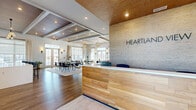 The Heartland clubhouse is a thoughtfully designed single-floor amenity space that blends contemporary aesthetics with warm, natural materials. Upon entry, a striking reception desk clad in light wood paneling greets visitors, set against a textured stone-look accent wall bearing the Heartland name. The open-concept main area seamlessly connects the kitchen and living room, anchored by wide-plank hardwood-style flooring and coffered ceilings with rich wood inlays and recessed lighting throughout.

The kitchen is outfitted with slate-blue cabinetry, white quartz countertops, stainless steel appliances, and a textured brick-style accent wall that adds warmth and character. An island provides additional counter space, while large windows flood the area with natural light and outdoor views.

The living area is a curated lounge space featuring a linear stone fireplace, a flat-screen television, bold original artwork, and statement ring-chandelier lighting. Plush seating arrangements and patterned rugs create distinct zones for relaxing and socializing. A sun-drenched reading nook with floor-to-ceiling windows offers a quieter retreat.

A private workstation with a built-in desk, large round mirror, and task lighting provides a functional workspace. A hallway connects the main areas to the full bathroom, completing this well-appointed communal facility.