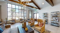 This single-story community space is defined by its striking architectural design, most notably the soaring vaulted ceilings accented by prominent exposed wood beams that run throughout the main lounge areas. Large, floor-to-ceiling grid-pane windows flood the interior with abundant natural light while framing views of palm trees and outdoor amenities, reinforcing a bright, coastal atmosphere.

The primary lounge area is generously proportioned, featuring polished large-format tile flooring, a statement multi-bulb chandelier, and a curated arrangement of upholstered wingback chairs and sofas in warm amber and neutral tones, accented with colorful throw pillows. A focal fireplace wall clad in herringbone-patterned tile anchors one end of the space, paired with a mounted television and built-in seating. Open metal shelving units displaying decorative objects line an adjacent wall, adding visual interest.

A separate gathering room features a substantial solid-wood communal dining table surrounded by leather and upholstered chairs, set against a wall of cabinetry with open shelving and multiple windows. Exposed ceiling beams and recessed lighting complete this functional, well-appointed area. Throughout, the design balances warm materials with clean, contemporary finishes to create an inviting and versatile environment.