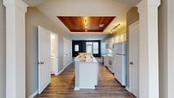 This two-bedroom, three-bathroom townhouse in Toledo, Ohio offers a well-organized two-floor layout designed for functional everyday living. The main floor makes an immediate impression in the kitchen, which features a striking coffered ceiling clad in rich wood paneling with integrated lighting — a clear architectural focal point. White cabinetry, speckled stone countertops, a center island with a sink, and white appliances complete the space. Durable hardwood-style flooring runs throughout the main level, connecting the kitchen seamlessly to the living room, which is accented by a deep blue feature wall and natural light from front-facing windows. A hallway with multiple doorways provides convenient access to a bathroom on the main floor. Upstairs, the two bedrooms are carpeted and bright, each featuring windows that bring in natural light and vaulted ceiling lines that lend an open, airy feel. The primary bedroom is generously sized, offering ample room for furnishings. The bathrooms are finished with speckled stone vanity countertops and include a soaking tub with tiled surround. The upper floor also includes a hallway connecting the bedrooms and bathrooms. A staircase links both levels, completing this practical and well-appointed townhouse layout.