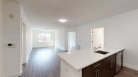 This single-floor residence showcases a clean, contemporary design with a cohesive aesthetic throughout. The open-concept layout seamlessly connects the kitchen and living area, creating a generous, airy feel enhanced by crisp white walls and rich hardwood-style flooring. The kitchen is thoughtfully designed with dark walnut-toned cabinetry, sleek white countertops, a kitchen island with an integrated sink, and a full suite of stainless steel appliances including a gas range, built-in microwave, dishwasher, and refrigerator. A mosaic tile backsplash adds a stylish accent to the cooking space. The living area is bright and open, benefiting from natural light streaming in through large multi-pane windows. Multiple bedrooms continue the clean, minimalist aesthetic, each featuring hardwood-style flooring, white walls, and windows that offer views of the surrounding neighborhood. The bathroom is elegantly finished with large-format light tile flooring, a dark wood vanity with white countertop, a wide mirror with overhead lighting, and a tub-shower combination framed by classic white subway tiles accented with a dark mosaic border. The overall ambiance is modern, fresh, and move-in ready.