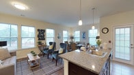 This single-floor residence is thoughtfully designed with an open-concept layout that seamlessly connects the kitchen, living, and dining areas into one cohesive, light-filled space. Multiple large windows throughout the main living area invite abundant natural light and offer outdoor views, creating an airy and welcoming atmosphere. The kitchen is well-appointed with rich dark cabinetry, granite countertops, a subway tile backsplash, and a full suite of stainless steel appliances. A central island with an undermount sink and pendant lighting above serves as both a functional workspace and a natural dividing point between the kitchen and living areas. The dining area comfortably accommodates a full-sized table and is surrounded by windows on multiple walls. A separate flex room provides additional living space ideal for a sitting room or den, featuring neutral carpet and soft, muted wall tones. The bedroom is generously sized with plush carpeting, a soothing color palette, and access to multiple closets. Two bathrooms complete the residence — one featuring a tiled walk-in shower, an extended vanity with dark cabinetry, and a large mirror, while the second is a more compact full bath finished with neutral tile flooring and cool gray walls.