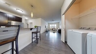 This 2-bedroom, 1-bathroom residence is thoughtfully designed to maximize both comfort and functionality across a single floor. The open-concept kitchen and living area serves as the heart of the home, featuring dark hardwood-style flooring that flows seamlessly throughout. The kitchen is well-equipped with rich dark cabinetry, granite countertops, a tile backsplash, and a full suite of stainless steel appliances, including a range, microwave, and refrigerator. A breakfast bar with pendant lighting provides casual seating and connects the kitchen to the main living space. The living room is bright and welcoming, with a large window that invites natural light and a ceiling fan overhead. Both bedrooms are comfortably sized, each featuring plush carpeting, a window with blinds, and ample space for a bed and desk — making them well-suited for rest or work. The shared bathroom is finished with dark cabinetry, granite countertops, a large mirror, and wood-look tile flooring, complemented by a full soaking tub and shower combination. A dedicated in-unit laundry room with a full-size washer and dryer adds everyday convenience. The overall aesthetic is clean, modern, and neutral, creating a versatile and move-in-ready living environment.
