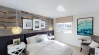 This thoughtfully designed single-floor space exudes a refined coastal aesthetic, blending natural textures with a clean, modern palette. The standout feature of the bedroom is a striking reclaimed wood accent wall, its weathered grey and warm brown tones adding rustic character and depth to the otherwise crisp white surroundings. A plush, dark upholstered bed anchors the room, dressed in bright white linens that enhance the light and airy feel. An eye-catching sculptural pendant light adds a touch of artistic elegance, while framed black-and-white coastal photography on the accent wall reinforces the nautical theme. Natural light pours in through a large window fitted with a woven roman shade, offering a picturesque view of a marina dotted with sailboats — a serene and distinctive backdrop that elevates the room's ambiance. Neutral carpet underfoot and white walls throughout keep the space feeling open and balanced. The overall atmosphere is calm, curated, and inviting, combining the warmth of natural materials with contemporary design sensibilities for a space that feels both stylish and comfortable.