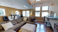 Located at 98 Oak Street in Lindenwold, New Jersey, this single-floor home offers a thoughtfully designed layout that balances comfort and functionality. The main living area immediately draws attention with its rich hardwood flooring and warm, earthy wall tones that create an inviting atmosphere throughout. A generously sized sectional sofa anchors the living space, complemented by an ottoman and area rug, making it ideal for relaxing or entertaining. Natural light floods the room through a series of large, white-trimmed double-hung windows, offering pleasant outdoor views and enhancing the open feel of the space. A distinctive circular accent window adds a unique architectural touch to the room's character. The elevated ceiling height adds a sense of volume, with upper-level windows providing additional light and visual interest. A built-in desk and shelving area along one wall offers a dedicated workspace seamlessly integrated into the living space. The overall ambiance of the home is warm and welcoming, with consistent finishes and thoughtful design details that contribute to a cohesive and comfortable living environment throughout.