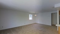 This single-floor space presents a clean, minimalist interior centered around an open living area. The room is finished with wall-to-wall beige carpeting that adds warmth underfoot, while crisp white walls and a smooth white ceiling create a bright, airy atmosphere. A single window allows natural light to filter into the space, illuminating the neutral tones throughout. A simple pendant light fixture hangs from the ceiling, providing a focal point and supplemental overhead lighting. The overall layout is open and versatile, offering a blank canvas that can accommodate a variety of furniture arrangements and design preferences. The absence of clutter and additional architectural features keeps the aesthetic straightforward and functional, making it well-suited for those looking to personalize the space to their own taste. The single-story design ensures easy accessibility throughout, and the understated finishes provide a neutral foundation that pairs well with a wide range of interior styles, from contemporary to traditional.