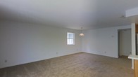 This single-floor space presents a clean, minimalist interior centered around an open living area. The room is finished with wall-to-wall beige carpeting that adds warmth underfoot, while crisp white walls and a smooth white ceiling create a bright, airy atmosphere. A single window allows natural light to filter into the space, illuminating the neutral tones throughout. A simple pendant light fixture hangs from the ceiling, providing a focal point and supplemental overhead lighting. The overall layout is open and versatile, offering a blank canvas that can accommodate a variety of furniture arrangements and design preferences. The absence of clutter and additional architectural features keeps the aesthetic straightforward and functional, making it well-suited for those looking to personalize the space to their own taste. The single-story design ensures easy accessibility throughout, and the understated finishes provide a neutral foundation that pairs well with a wide range of interior styles, from contemporary to traditional.