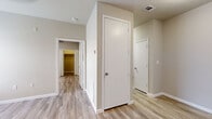 This single-floor unit offers a thoughtfully organized layout designed to maximize functionality within a compact footprint. Light wood-look luxury vinyl flooring runs consistently throughout, lending a cohesive and clean aesthetic to every room. Neutral warm-gray walls complement the pale flooring, creating a bright and airy ambiance across the entire space.

The galley-style kitchen is well-appointed with white shaker cabinetry extending from floor to ceiling on both sides, providing ample storage. A stainless steel double-basin sink, white countertops, an under-cabinet range hood, and a black electric range complete the kitchen's practical setup.

Multiple rooms branch off a central hallway, each featuring ceiling fan fixtures and windows fitted with blinds that allow natural light to filter in. One room includes a walk-in closet with built-in shelving, while another features a standard reach-in closet. The rooms offer versatile potential for sleeping or living arrangements.

The bathroom includes a full tub-shower combination with a built-in niche, a toilet, and a vanity with a white countertop and modern hardware. The entryway opens into a spacious living area, grounded by the same consistent flooring and neutral tones that define the rest of the unit.