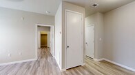 This single-floor unit offers a thoughtfully organized layout designed to maximize functionality within a compact footprint. Light wood-look luxury vinyl flooring runs consistently throughout, lending a cohesive and clean aesthetic to every room. Neutral warm-gray walls complement the pale flooring, creating a bright and airy ambiance across the entire space.

The galley-style kitchen is well-appointed with white shaker cabinetry extending from floor to ceiling on both sides, providing ample storage. A stainless steel double-basin sink, white countertops, an under-cabinet range hood, and a black electric range complete the kitchen's practical setup.

Multiple rooms branch off a central hallway, each featuring ceiling fan fixtures and windows fitted with blinds that allow natural light to filter in. One room includes a walk-in closet with built-in shelving, while another features a standard reach-in closet. The rooms offer versatile potential for sleeping or living arrangements.

The bathroom includes a full tub-shower combination with a built-in niche, a toilet, and a vanity with a white countertop and modern hardware. The entryway opens into a spacious living area, grounded by the same consistent flooring and neutral tones that define the rest of the unit.