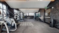 This contemporary fitness center is designed with both form and function in mind, offering a comprehensive workout environment across a single open floor. The space features a striking industrial-modern aesthetic, highlighted by textured geometric wall panels in deep charcoal tones that lend a bold, sophisticated atmosphere throughout.

Natural light flows generously into the facility through expansive floor-to-ceiling windows along multiple walls, as well as overhead skylights that illuminate the interior and provide open views of the surrounding cityscape. The large-format gray tile flooring provides a durable, clean surface suited for high-traffic fitness use.

The cardio zone is well-appointed with stair climbers, elliptical machines, and treadmills positioned along the windowed walls to take advantage of the natural light and outdoor views. The strength training area includes a multi-station cable machine, adjustable benches, a dedicated free weights rack with a full dumbbell set, a punching bag, and additional resistance equipment.

Mirrored wall panels expand the sense of space and support proper form during training. A water fountain station with dual dispensers is conveniently located within the facility. Recessed and linear LED lighting complements the natural light, ensuring consistent illumination throughout. The overall ambiance is energetic yet refined, making it a functional and visually impressive fitness facility.
