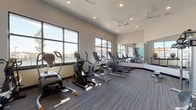 The fitness center at Haven on Tucker is a thoughtfully designed, single-floor space built to support a comprehensive workout experience. The room is anchored by large, floor-to-ceiling windows along multiple walls, allowing abundant natural light to pour in and offering views of the surrounding outdoor amenity area, including a pool and neighboring community. Ceiling fans and recessed lighting ensure the space remains comfortable and well-lit throughout the day. A standout design feature is the bold geometric accent wall, rendered in teal, burgundy, and gray tones, which adds a vibrant, energetic character to the space. Full-length mirrors are mounted along one wall, enhancing the sense of openness and providing a practical resource for form-checking during workouts. The flooring consists of durable, striped commercial-grade carpet tiles suited for high-traffic fitness use. Equipment is well-organized and includes multiple treadmills, elliptical trainers, stationary and air bikes, a stair climber, a cable weight machine, a dumbbell rack, and a rowing machine, offering options for both cardio and strength training. The layout provides generous spacing between machines, allowing for comfortable movement throughout the room. Overall, the fitness center combines functional design with an inviting, modern aesthetic.