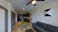 This 2-bedroom, 2-bathroom single-floor residence offers an open-concept design that seamlessly connects the living, dining, and kitchen areas. The living space is anchored by a ceiling fan with integrated lighting and features smooth, light-toned hardwood-style flooring that runs throughout the main area, contributing to a clean and cohesive aesthetic.

The kitchen is outfitted with dark cabinetry, black appliances including a full-size refrigerator, microwave, and range, as well as a sink with an adjacent countertop workspace. Track lighting above the kitchen provides focused illumination for food preparation. A compact dining area sits adjacent to the kitchen, furnished with a round table and modern green chairs, making efficient use of the transitional space.

The living area provides comfortable seating and connects naturally to the rest of the unit. Neutral white walls throughout keep the space feeling bright and open. Doorways visible from the main area lead to the bedrooms and bathrooms, offering a practical and functional layout. Overall, the unit presents a modern, well-organized living environment suited for everyday comfort.
