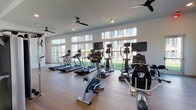 This fitness center is designed to support a comprehensive workout experience across multiple functional zones. The main cardio area is lined with floor-to-ceiling windows that frame views of a landscaped outdoor courtyard, flooding the space with natural light. Multiple treadmills, ellipticals, upright bikes, and a recumbent bike are arranged to take full advantage of the view, each unit equipped with an individual display screen. Ceiling fans are mounted overhead, maintaining airflow throughout the open layout, while light hardwood flooring runs consistently across all areas, contributing to a clean, modern aesthetic.

A strength training zone features a multi-station cable machine, free weights, a weight rack, and additional functional fitness equipment, providing options for resistance and circuit-based workouts. Large windows in this area continue the theme of natural light and outdoor connectivity.

A dedicated studio room offers a more intimate setting, finished with smooth white walls, recessed lighting, a wall-mounted television, a mirror, and specialty equipment including a reformer-style training system. This room is well-suited for focused, low-impact training or stretching routines. Together, the zones create a well-rounded, modern fitness facility with a bright, airy atmosphere throughout.