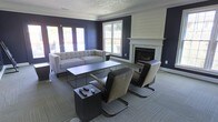 This single-floor space presents a refined and contemporary aesthetic, centered around a well-appointed lounge area that balances modern design with classic architectural details. The room is anchored by a striking white shiplap fireplace surround, which serves as a focal point against the bold navy-blue accent walls, creating a dramatic yet inviting contrast. Crown molding adds a traditional elegance to the space, while a decorative ceiling tile pattern provides an additional layer of visual interest overhead. Natural light flows generously into the room through a set of large windows and French doors, offering views of the outdoor surroundings and enhancing the open, airy feel of the interior. The lounge seating arrangement features a tufted light gray sofa paired with two contemporary cantilever-style accent chairs in warm earth tones, all centered around a sleek, low-profile rectangular coffee table. A compact side table with integrated storage completes the seating grouping. The flooring is finished in a patterned low-pile carpet in neutral gray tones, grounding the space and adding texture underfoot. Overall, the space conveys a sophisticated and comfortable ambiance suited for relaxed gathering or quiet lounging.