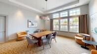 Located at 243 Steele Rd in West Hartford, CT, this refined dining room offers a thoughtfully designed space that blends modern sophistication with mid-century charm. The room is anchored by a rich walnut rectangular dining table surrounded by upholstered grey chairs, providing comfortable seating for gatherings. A striking elongated pendant light in brushed metallic tones hangs above the table, complementing the tray ceiling fitted with recessed lighting that adds warmth and depth to the space. A patterned area rug in warm gold and neutral tones grounds the room and adds texture underfoot. Large multi-pane windows line one wall, inviting generous natural light and offering pleasant views of the surrounding neighborhood. A mid-century credenza along the opposite wall is adorned with a triptych of abstract watercolor artwork in cool teal and green hues, lending an artistic focal point. Accent seating in the form of curved amber leather lounge chairs with small side tables creates an inviting conversational nook, adding versatility to the space. Soft grey walls and crisp white trim throughout create a clean, airy backdrop that ties the entire room together with understated elegance.