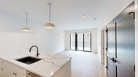 This thoughtfully designed one-bedroom, one-bathroom unit offers a clean, contemporary aesthetic across its single floor. The open-concept kitchen and living area serves as the heart of the unit, anchored by a striking oversized waterfall-edge quartz island with an undermount sink and matte black faucet. The kitchen is outfitted with dark cabinetry contrasted by white upper cabinets, a linear tile backsplash, stainless steel appliances including a French door refrigerator, microwave, and range, and pendant lighting overhead. Light-toned hardwood-style flooring flows seamlessly throughout, complementing the crisp white walls and recessed lighting. The living area opens directly to an outdoor space through full-width black-framed sliding glass doors, flooding the interior with natural light. The bedroom is a comfortable, well-proportioned room featuring its own window with an exterior view and access to a dedicated closet. The bathroom offers a clean, minimalist design with marble-style tile flooring, a single undermount vanity with white countertop, and a full-size mirror. Bold black interior doors provide a consistent modern accent throughout the unit.