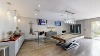 Located at 100 Myrtle St in Longwood, Florida, this single-floor game and recreation room is designed with a contemporary aesthetic and a strong focus on social entertainment. The space is anchored by a centerpiece pool table with a neutral felt surface, set beneath a sleek rectangular pendant light fixture that provides focused, stylish illumination. Multiple wall-mounted flat-screen TVs are positioned throughout the room, ensuring excellent viewing angles from nearly every corner.

The flooring features large-format stone-look tile that adds texture and warmth to the space. Bar-height round tables paired with modern black stools are distributed across the room, offering casual seating arrangements ideal for gathering and conversation. A built-in counter along one wall provides additional surface space, complemented by a wall-mounted cue rack for convenient storage of billiard equipment. Decorative artwork, including bold canvas pieces and a colorful abstract painting, add visual interest to the neutral gray walls.

One of the room's standout features is its large sliding glass doors that flood the interior with natural light and open directly to an outdoor area complete with a pool and lush tropical landscaping, seamlessly blending indoor and outdoor living. The overall ambiance is modern, vibrant, and well-suited for recreational use and community gatherings.