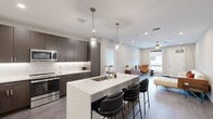 This contemporary single-floor residence showcases a thoughtfully designed open-concept layout that seamlessly connects the kitchen, dining, and living areas. The gourmet kitchen is a standout feature, outfitted with dark wood cabinetry, crisp white quartz countertops, stainless steel appliances including a refrigerator, microwave, and range, and a large central island with an undermount sink and seating for multiple bar stools. Elegant pendant lighting hangs above the island, adding a refined touch to the space. The living area is bright and welcoming, featuring wood-style flooring, recessed lighting, a ceiling fan, and large windows that invite natural light and outdoor views. The bedrooms are carpeted for comfort, with generously sized windows fitted with roller shades offering serene outdoor views. The bathrooms carry the same modern aesthetic, featuring dark wood vanities, white quartz countertops, and wood-look tile flooring. Neutral tones in warm greige and white are carried consistently throughout the unit, creating a clean, cohesive ambiance. The overall design reflects a modern sensibility with high-quality finishes and a functional, flowing floor plan.