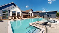 Located at 5617 Oakley Industrial Blvd in South Fulton, Georgia, this outdoor amenity space presents a well-appointed resort-style pool area designed for both relaxation and social gatherings. The centerpiece is a generously sized swimming pool with crystal-clear blue water, entry steps, and stainless steel handrails, complemented by a separate, smaller wading or spa pool nearby. The surrounding deck is finished in a light-toned concrete surface that provides ample space for lounging and entertaining.

The pool deck is outfitted with a variety of seating options, including cushioned chaise lounges, wicker lounge chairs, and grouped conversation sets positioned beneath striped market umbrellas that offer shade throughout the day. A pergola structure adds an architectural focal point to the seating area, enhancing the outdoor ambiance. Decorative sculptural elements and potted greenery punctuate the space, contributing to its polished aesthetic.

The area is enclosed by a black metal fence and is framed by mature trees that provide a natural backdrop. Multi-story residential buildings in navy blue, white, and earth-tone exteriors surround the pool, creating a cohesive community setting. The space enjoys abundant natural light under open Georgia skies, making it a vibrant outdoor destination.