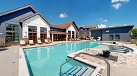 Located at 5617 Oakley Industrial Blvd in South Fulton, Georgia, this outdoor amenity space presents a well-appointed resort-style pool area designed for both relaxation and social gatherings. The centerpiece is a generously sized swimming pool with crystal-clear blue water, entry steps, and stainless steel handrails, complemented by a separate, smaller wading or spa pool nearby. The surrounding deck is finished in a light-toned concrete surface that provides ample space for lounging and entertaining.

The pool deck is outfitted with a variety of seating options, including cushioned chaise lounges, wicker lounge chairs, and grouped conversation sets positioned beneath striped market umbrellas that offer shade throughout the day. A pergola structure adds an architectural focal point to the seating area, enhancing the outdoor ambiance. Decorative sculptural elements and potted greenery punctuate the space, contributing to its polished aesthetic.

The area is enclosed by a black metal fence and is framed by mature trees that provide a natural backdrop. Multi-story residential buildings in navy blue, white, and earth-tone exteriors surround the pool, creating a cohesive community setting. The space enjoys abundant natural light under open Georgia skies, making it a vibrant outdoor destination.