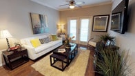 This single-story space centers around a thoughtfully designed living area that balances comfort and style. The room features rich hardwood flooring that anchors the space and contributes to a warm, welcoming ambiance throughout. Crown molding along the ceiling adds a refined architectural detail, while a ceiling fan with integrated lighting provides both practicality and a classic finishing touch.

A defining feature of the living area is the set of white French doors, fitted with horizontal blinds, which flood the room with natural light and provide direct access to an outdoor area. The doors serve as a natural focal point, enhancing the sense of openness and connectivity to the exterior.

The living space is generously proportioned, comfortably accommodating a full-sized sofa, accent chairs, and a nesting coffee table arrangement atop a plush area rug. Neutral wall tones in warm gray provide a versatile backdrop that complements the colorful decorative accents, including framed artwork and vibrant throw pillows. The overall ambiance is one of casual sophistication — a bright, livable space designed for everyday comfort.