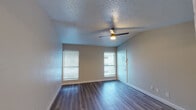 This single-floor unit offers a clean, functional layout designed for comfortable everyday living. The main living area is bright and open, featuring durable wood-look plank flooring, neutral-toned walls, and a ceiling fan with integrated lighting. Two windows dressed with horizontal blinds allow natural light to fill the space while maintaining privacy. The living area flows seamlessly into the kitchen, which is defined by a peninsula counter with a dark countertop surface, white upper cabinetry, and stainless steel appliances including a refrigerator and range. Adjacent to the kitchen, a dedicated laundry area provides added convenience. The bedroom continues the wood-look flooring and offers a window with blinds, creating a calm and functional retreat. A ceiling fan is also present in this room. The full bathroom is well-appointed with a white vanity featuring a single undermount sink, dark countertop, a framed mirror, and a well-lit vanity bar above. A combined tub and shower with a rainfall-style showerhead completes the space. Throughout, textured popcorn ceilings and clean white baseboards contribute to a consistent and practical interior aesthetic.