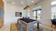 This inviting game room blends modern farmhouse aesthetics with a well-curated selection of recreational amenities. The space is anchored by a substantial pool table with a black felt surface and a weathered wood frame, positioned atop a richly patterned area rug that adds warmth and character to the whitewashed hardwood floors below. A striking cluster of three antique brass pendant lights hangs directly above the table, providing focused illumination while serving as a bold design statement.

One wall features a natural wood plank accent panel that houses a wall-mounted flat-screen television alongside a built-in billiard cue rack, keeping equipment both accessible and neatly organized. Opposite this, a large framed wall game board adds a playful, decorative touch. A cozy lounge nook in the corner offers a round bistro table flanked by two sleek leather chairs — an ideal spot for chess or card games.

Soaring ceilings with a white-painted shiplap finish amplify the sense of openness, while oversized windows with transoms along two walls flood the room with natural light and offer views of the surrounding outdoor landscape. Glass-paned double doors provide direct access to an exterior patio. The overall ambiance is bright, relaxed, and well-appointed for social entertainment.