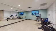 This contemporary fitness studio is thoughtfully designed to accommodate a wide range of exercise disciplines within a single, open space. The room features light wood-look flooring throughout, creating a clean and inviting atmosphere complemented by soft sage-green accent walls and recessed lighting overhead.

A floor-to-ceiling mirror spans one full wall, paired with dual-level wooden ballet barres — ideal for barre classes, stretching, and dance-inspired workouts. Mounted flat-screen televisions are positioned along the walls, enabling guided virtual workout sessions and on-demand class streaming directly within the studio.

The cycling area houses multiple stationary bikes equipped with individual screens, supporting interactive and instructor-led cycling programs. A dedicated strength and functional training zone features a well-stocked equipment rack displaying kettlebells, medicine balls, resistance tools, a TRX suspension trainer, and a heavy punching bag, alongside plyo boxes for high-intensity training.

A striking line-art mural of floral and botanical motifs decorates one wall, adding an artistic and calming visual element to the energetic environment. Built-in storage cabinetry in a warm wood finish provides organized space for equipment and accessories. The studio is completed by a private entry door, ensuring a self-contained and focused workout environment suitable for individual training and group fitness alike.