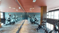 This fitness center spans two floors and is designed to deliver a motivating and functional workout environment. The space is immediately distinguished by its bold interior color scheme, featuring deep teal walls contrasted with a warm coral-toned ceiling, creating a vibrant and energizing atmosphere. Floor-to-ceiling mirrors run along one wall, amplifying the sense of space and allowing members to monitor their form during exercise. The flooring features a patterned, light-toned carpet that adds a contemporary touch while providing underfoot comfort. The cardio zone is well-equipped with a row of elliptical trainers and stationary cycling bikes, arranged to maximize the available floor space. Recessed lighting throughout ensures the area is well-lit, while the open layout promotes easy movement between stations. A warm wood accent column adds an architectural detail that softens the industrial feel of the equipment. The two-floor layout suggests additional workout areas, potentially housing strength training equipment, free weights, or dedicated studio space on a separate level. Overall, the fitness center balances functional design with thoughtful aesthetics, making it a welcoming space for a wide range of fitness activities.