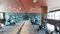 This fitness center spans two floors and is designed to deliver a motivating and functional workout environment. The space is immediately distinguished by its bold interior color scheme, featuring deep teal walls contrasted with a warm coral-toned ceiling, creating a vibrant and energizing atmosphere. Floor-to-ceiling mirrors run along one wall, amplifying the sense of space and allowing members to monitor their form during exercise. The flooring features a patterned, light-toned carpet that adds a contemporary touch while providing underfoot comfort. The cardio zone is well-equipped with a row of elliptical trainers and stationary cycling bikes, arranged to maximize the available floor space. Recessed lighting throughout ensures the area is well-lit, while the open layout promotes easy movement between stations. A warm wood accent column adds an architectural detail that softens the industrial feel of the equipment. The two-floor layout suggests additional workout areas, potentially housing strength training equipment, free weights, or dedicated studio space on a separate level. Overall, the fitness center balances functional design with thoughtful aesthetics, making it a welcoming space for a wide range of fitness activities.