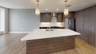 This single-floor residence showcases a thoughtfully designed contemporary interior with a cohesive blend of warm wood tones, clean white surfaces, and bold accent colors throughout. The open-concept kitchen serves as a central highlight, featuring a generously sized island with a crisp white quartz countertop, an undermount sink, and pendant lighting overhead. Dark wood-grain cabinetry lines the walls alongside stainless steel appliances, including a range with a wall-mounted hood and a full-size refrigerator, all complemented by a neutral tile backsplash.

The living areas are finished with hardwood-style flooring that flows seamlessly throughout the space, lending warmth and visual continuity. A bedroom features a striking teal accent wall paired with a ceiling fan and hardwood floors, offering a calm, restful atmosphere.

The bathroom is well-appointed, showcasing a large glass-enclosed walk-in shower clad in earthy vertical tile, wood-toned vanity cabinetry with white countertops, and light wood-look tile flooring. A teal ceiling detail adds a design-forward touch.

The living area is defined by an impressive wall of oversized floor-to-ceiling windows that frame sweeping, elevated panoramic city views, flooding the space with abundant natural light and providing a dramatic focal point for the entire residence.