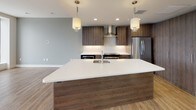 This single-floor residence showcases a thoughtfully designed contemporary interior with a cohesive blend of warm wood tones, clean white surfaces, and bold accent colors throughout. The open-concept kitchen serves as a central highlight, featuring a generously sized island with a crisp white quartz countertop, an undermount sink, and pendant lighting overhead. Dark wood-grain cabinetry lines the walls alongside stainless steel appliances, including a range with a wall-mounted hood and a full-size refrigerator, all complemented by a neutral tile backsplash.

The living areas are finished with hardwood-style flooring that flows seamlessly throughout the space, lending warmth and visual continuity. A bedroom features a striking teal accent wall paired with a ceiling fan and hardwood floors, offering a calm, restful atmosphere.

The bathroom is well-appointed, showcasing a large glass-enclosed walk-in shower clad in earthy vertical tile, wood-toned vanity cabinetry with white countertops, and light wood-look tile flooring. A teal ceiling detail adds a design-forward touch.

The living area is defined by an impressive wall of oversized floor-to-ceiling windows that frame sweeping, elevated panoramic city views, flooding the space with abundant natural light and providing a dramatic focal point for the entire residence.