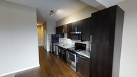 This single-floor space showcases a contemporary design anchored by a well-appointed kitchen. The kitchen features floor-to-ceiling dark espresso cabinetry that provides generous storage, complemented by light granite countertops that create a striking contrast. Stainless steel appliances, including an electric range, built-in microwave, and refrigerator, lend a polished, modern aesthetic. A stainless steel sink with a sleek faucet is integrated into the countertop, adding both function and style. Rich, dark hardwood flooring runs continuously throughout the space, enhancing the warm yet sophisticated atmosphere. Track lighting above the kitchen provides focused illumination, highlighting the cabinetry and countertops effectively. A hallway visible beyond the kitchen suggests additional rooms within the unit, with doors hinting at closet or storage space. The neutral grey walls throughout maintain a clean, understated backdrop that allows the dark cabinetry and flooring to remain the focal points. Overall, the space presents a cohesive, modern interior with a thoughtful balance of dark and light tones.