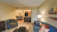This single-floor residence features an open-concept layout that seamlessly connects the living room, dining area, and kitchen, creating a cohesive and functional flow. The living room is furnished with blue upholstered seating accented by decorative pillows in warm orange and cream tones, complemented by a dark coffee table and a standing floor lamp that casts a warm ambient glow. Neutral carpet flooring anchors the living area, transitioning into hardwood-style flooring in the dining and kitchen zones. The dining area accommodates a dark wood table with matching chairs and is adorned with a framed piece of wall art, adding a touch of color to the warm beige walls. The kitchen features medium-tone wood cabinetry, dark countertops, and black appliances, including a full-size refrigerator. A breakfast bar-style counter separates the kitchen from the dining space, offering both functionality and visual definition. The warm yellow-beige wall color carries throughout, contributing to an inviting and cohesive ambiance. A white interior door near the living area provides access to an additional room, suggesting further private living space beyond the open common areas.