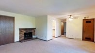 This single-floor space offers an open and functional layout with several notable features. The main living area is anchored by a stone-faced fireplace with a raised brick hearth, providing a classic focal point and a cozy atmosphere. The flooring throughout is wall-to-wall carpet in a neutral beige tone, contributing to a warm and understated aesthetic. The walls are painted in soft, light hues, and the textured ceiling adds subtle visual interest overhead.

A partial dividing wall separates the living area from an adjoining space, which opens into the kitchen. The kitchen is visible through the passageway and features light wood cabinetry, offering practical storage. A ceiling fan is installed in the adjacent area, providing both lighting and air circulation.

A hallway leads further into the home, with what appears to be additional rooms accessible beyond. Dark wood-toned doors are visible along the corridor, maintaining a consistent design language throughout. The overall ambiance of the space is straightforward and livable, with a neutral palette that provides a versatile foundation for a variety of interior styles.