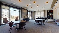 This elegantly designed single-floor amenity space offers a seamless blend of collaborative and private environments suited for work, meetings, and social gatherings. The main lounge area is generously proportioned, featuring patterned carpet with gold accents, a crisp white ceiling adorned with brass flush-mount and pendant light fixtures that lend a warm, upscale ambiance. Multiple seating arrangements—ranging from large rectangular tables with sleek dark chairs to intimate groupings with velvet accent seating—provide flexible use for various occasions. Floor-to-ceiling windows line the perimeter, flooding the space with abundant natural light and offering open views of the surrounding urban landscape. A navy blue credenza anchors the reception area, topped with white quartz and flanked by tall decorative vases. A lounge nook near the entrance features high-back swivel chairs positioned to take advantage of the panoramic views. A dedicated private meeting room, enclosed in glass, offers a more intimate setting with plush seating and curated artwork. Throughout the space, bold abstract murals, sculptural light installations, and carefully selected wall art contribute to a polished, gallery-like atmosphere. The overall design balances modern sophistication with functional versatility.