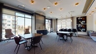 This elegantly designed single-floor amenity space offers a seamless blend of collaborative and private environments suited for work, meetings, and social gatherings. The main lounge area is generously proportioned, featuring patterned carpet with gold accents, a crisp white ceiling adorned with brass flush-mount and pendant light fixtures that lend a warm, upscale ambiance. Multiple seating arrangements—ranging from large rectangular tables with sleek dark chairs to intimate groupings with velvet accent seating—provide flexible use for various occasions. Floor-to-ceiling windows line the perimeter, flooding the space with abundant natural light and offering open views of the surrounding urban landscape. A navy blue credenza anchors the reception area, topped with white quartz and flanked by tall decorative vases. A lounge nook near the entrance features high-back swivel chairs positioned to take advantage of the panoramic views. A dedicated private meeting room, enclosed in glass, offers a more intimate setting with plush seating and curated artwork. Throughout the space, bold abstract murals, sculptural light installations, and carefully selected wall art contribute to a polished, gallery-like atmosphere. The overall design balances modern sophistication with functional versatility.