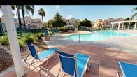 Located in Canyon Country, CA, this outdoor community amenity space centers around a generously sized freeform swimming pool with crystal-clear blue water, designed to serve as a central gathering and recreation hub. The pool deck is finished with terracotta-toned tile pavers and is outfitted with multiple blue lounge chairs, offering ample space for sunbathing and relaxation under the Southern California sun. A white pergola structure provides a shaded retreat adjacent to the pool, beneath which residents can enjoy outdoor seating while remaining sheltered from direct sunlight. A gas BBQ grill is conveniently positioned nearby, making the area well-suited for outdoor entertaining. Mature palm trees and a variety of drought-tolerant shrubs and flowering plants are thoughtfully landscaped throughout the space, adding lush greenery and visual appeal. The area is enclosed by a green metal security fence, ensuring safety and privacy. The surrounding residential buildings feature a classic Spanish-inspired architectural style with warm stucco exteriors, tiled rooflines, and arched windows, creating a cohesive and inviting community atmosphere. The backdrop of the Santa Clarita Valley hillsides further enhances the scenic character of this well-maintained outdoor amenity space.