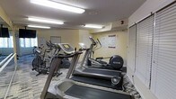 This fitness center is a well-appointed single-floor exercise space designed to support a range of cardio-focused workouts. The room features multiple Precor-brand machines, including treadmills, ellipticals, and stationary bikes, arranged to make efficient use of the available floor space. The walls are painted in a soft yellow tone, contributing to a bright and energizing atmosphere, while the textured ceiling houses fluorescent lighting that ensures the room is well-lit throughout. Informational posters and workout guides are mounted on the walls, offering users guidance and reference during their sessions. The flooring features a patterned carpet, and a louvered vent panel is visible along one wall, suggesting adequate ventilation for the space. The layout is straightforward and functional, positioning equipment in a way that allows users to move comfortably between machines. While compact in scale, the fitness center provides a practical and accessible option for cardiovascular exercise, making it a convenient amenity for residents or guests seeking to maintain an active routine without leaving the building.