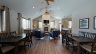 This single-story community clubhouse is thoughtfully designed to foster comfort and connection. The centerpiece of the space is a spacious great room boasting soaring vaulted ceilings with ceiling fans, rich hardwood-style flooring, and an abundance of natural light streaming through multiple windows dressed with patterned curtains and wood-trimmed blinds. A tile-surround gas fireplace with a classic wood mantel serves as a striking focal point, complemented by a wall-mounted flat-screen television above. The seating area is furnished with plush sofas and accent chairs, creating an inviting atmosphere for socializing or unwinding.

Multiple dining tables with dark wood chairs are arranged throughout the open layout, accommodating gatherings of various sizes. A sliding glass door in the dining area provides access to an outdoor patio, further enhancing the sense of openness. The full kitchen is equipped with warm honey-toned cabinetry, granite-look countertops, black appliances including a range and microwave, and tile flooring — offering practical amenities for events and everyday use.

Beyond the main living area, glass-paneled interior doors lead to a dedicated fitness room and a hallway connecting additional amenities. The overall ambiance is warm, functional, and welcoming, making this clubhouse an excellent community hub.