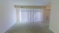 This single-floor space features a generously sized main living area characterized by its simple, functional design. The room is finished with neutral-toned wall-to-wall carpeting that extends throughout, providing a soft underfoot feel and a cohesive, understated aesthetic. The walls are painted in a light, off-white tone, contributing to a bright and airy atmosphere.

A standout feature of the room is the expansive bank of vertical blinds that span nearly the entire width of one wall, suggesting the presence of large sliding glass doors or an oversized window behind them. This installation allows for significant natural light to filter through when the blinds are open, and offers privacy and light control when closed. The structural soffit above the window treatment adds a defined architectural element to the room.

Ceiling-mounted HVAC vents are visible, indicating a central air and heating system integrated throughout the space. The room's open layout offers flexible arrangement possibilities, making it well-suited for a variety of living configurations. Overall, the space presents a clean, neutral canvas ready for personalization.