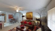 This single-floor space centers around a comfortable living room designed with a warm and inviting aesthetic. The room features a textured ceiling with a ceiling fan and light fixture, providing both airflow and ambient illumination. Soft, neutral-toned carpet runs throughout, contributing to a cozy atmosphere.

The living area is anchored by a plush, dark upholstered sofa adorned with rich red and patterned accent pillows, complemented by a solid wood coffee table with a decorative red bowl as a centerpiece. A bold landscape painting in warm amber and brown tones serves as a focal point on the main wall, while tall decorative vases with branching accents flank the seating area, adding height and visual interest.

Warm accent lighting from floor and table lamps enhances the room's inviting ambiance. An open sightline leads to a dining area in the background, where a round table and chairs are visible beneath additional lighting, suggesting a functional and connected layout. The space flows naturally between living and dining areas, making it well-suited for both relaxed everyday living and casual entertaining.