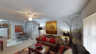 This single-floor space centers around a comfortable living room designed with a warm and inviting aesthetic. The room features a textured ceiling with a ceiling fan and light fixture, providing both airflow and ambient illumination. Soft, neutral-toned carpet runs throughout, contributing to a cozy atmosphere.

The living area is anchored by a plush, dark upholstered sofa adorned with rich red and patterned accent pillows, complemented by a solid wood coffee table with a decorative red bowl as a centerpiece. A bold landscape painting in warm amber and brown tones serves as a focal point on the main wall, while tall decorative vases with branching accents flank the seating area, adding height and visual interest.

Warm accent lighting from floor and table lamps enhances the room's inviting ambiance. An open sightline leads to a dining area in the background, where a round table and chairs are visible beneath additional lighting, suggesting a functional and connected layout. The space flows naturally between living and dining areas, making it well-suited for both relaxed everyday living and casual entertaining.