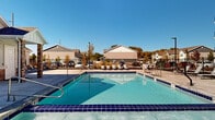This outdoor amenity space centers around a generously sized rectangular swimming pool with striking cobalt blue tile accents and crystal-clear turquoise water. The pool is complemented by an adjacent hot tub/spa, offering a relaxing hydrotherapy option alongside the main swimming area. Both are finished with matching blue tile detailing that adds a clean, polished aesthetic throughout.

Surrounding the pool is a broad concrete deck that provides ample space for lounging and socializing. Striped and solid lounge chairs are arranged along multiple sides of the deck, inviting residents or guests to relax in the open air. The entire area is enclosed by a black iron fence, providing both security and an unobstructed view of the surrounding landscape, which includes neighboring residential structures and mature trees that add seasonal color.

An adjacent single-story building — finished in a neutral palette with brick accents and a pitched roof — appears to serve as a poolside amenity or facility structure, providing convenient access to restrooms or additional services. Backdrop views include mountain silhouettes visible on the horizon, enhancing the outdoor atmosphere. The space is bathed in abundant natural sunlight, making it a bright, inviting environment ideal for outdoor recreation and leisure.