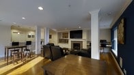 This well-appointed single-floor space is designed with both comfort and functionality in mind. The main living area serves as the heart of the space, anchored by a elegant fireplace with a mosaic tile surround and a classic white mantle. Above the fireplace, a built-in media niche houses a flat-screen television, while a cozy window seat with cushions and decorative pillows adds a charming architectural touch beneath. Rich hardwood flooring extends throughout the space, complementing the warm, inviting atmosphere. Distinctive white columns are positioned throughout, adding a sense of structure and architectural character without compromising the open feel of the layout. Dark leather seating is arranged thoughtfully around a wooden coffee table, creating an ideal gathering area. Recessed lighting illuminates the space evenly from the smooth white ceiling above. Adjacent areas are visible beyond the main living space, including what appears to be a dining or workspace area and an additional room or workspace to the right, suggesting a versatile, multi-functional layout. Colorful artwork on the walls adds visual interest, contributing to the polished and welcoming ambiance of the overall space.