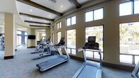 The fitness center at 11921 Carmel Creek Road in San Diego is a well-appointed, single-floor facility designed to support an active lifestyle. The space makes a strong first impression with its dramatic ceiling height, accentuated by dark exposed wood beams that add a refined architectural character to the room. Recessed lighting is strategically placed throughout, ensuring the space is well-lit at all times.

One of the most notable features is the expansive wall of large, floor-to-ceiling windows that line the exterior-facing side of the room. These windows flood the interior with natural light and offer open views of the surrounding outdoor courtyard and neighboring buildings, creating an airy and inviting atmosphere during workouts.

The facility is outfitted with a selection of modern cardio equipment, including multiple treadmills and elliptical machines, all arranged to take advantage of the natural light and views. A wall-mounted television is also present, providing entertainment options for users. The flooring is covered in a durable, patterned carpet suited for a fitness environment.

The warm, neutral-toned walls complement the overall design, and an open archway connects to an adjacent area, adding to the sense of spaciousness. Overall, this fitness center combines functional amenities with thoughtful design elements.
