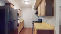 This single-floor residence offers a practical and comfortable layout designed for everyday living. The galley-style kitchen is efficiently arranged with light wood cabinetry lining both sides, butcher block-style countertops, and black appliances including a full-size refrigerator and an electric range. Hardwood-style flooring runs throughout the kitchen, adding warmth and easy maintenance to the space.

The living area is generously proportioned, featuring plush carpeting, a ceiling fan with light fixture, and open sightlines that create a welcoming atmosphere. An adjacent closet provides convenient storage access directly from the main living space. The transition between the carpeted and hardwood-style flooring areas delineates the living and kitchen zones naturally.

The bedroom is a well-sized, comfortable room with soft carpeting underfoot, a ceiling fan, and a window dressed with blinds that allows natural light to filter in while maintaining privacy. The view to the outdoors adds a pleasant connection to the surrounding environment.

The bathroom features a wide vanity with warm wood cabinetry, a single-basin sink with a light countertop, and a large framed mirror topped with a Hollywood-style light bar, providing ample illumination for daily routines. The hardwood-style flooring continues into the bathroom, maintaining a cohesive aesthetic throughout the unit.