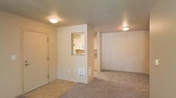This single-floor residential space offers a practical and well-organized layout designed for comfortable everyday living. The main living area is carpeted in a neutral tone, providing a warm and inviting atmosphere that flows seamlessly throughout the space. Flush-mount ceiling fixtures provide consistent overhead lighting, keeping the interior bright and functional.

A defining architectural feature is the pass-through window cut into a partial dividing wall, which connects the living area to the kitchen. This detail enhances the open feel of the space while maintaining a sense of separation between the two areas. The kitchen, visible through the pass-through, features light-colored cabinetry and white appliances, contributing to a clean and tidy aesthetic.

The entryway is accessible through a wood-toned door, offering a clear transition point into the living space. A wall-mounted floor vent provides in-unit climate control. The combination of tile flooring near the entry and plush carpet in the living and adjoining areas adds both practicality and comfort underfoot. Neutral beige wall tones throughout create a cohesive, understated palette that is easy to personalize. Overall, the space presents a straightforward, move-in-ready environment with all the essential elements for day-to-day living.